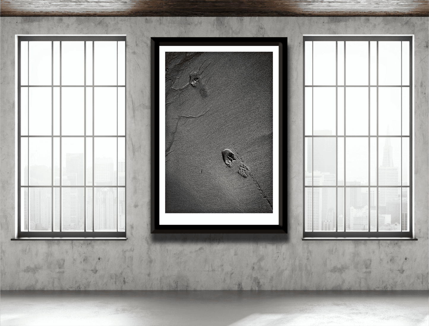 Large framed Traces #1 black and white sand photography with footprints, displayed in an industrial-style room with large windows.