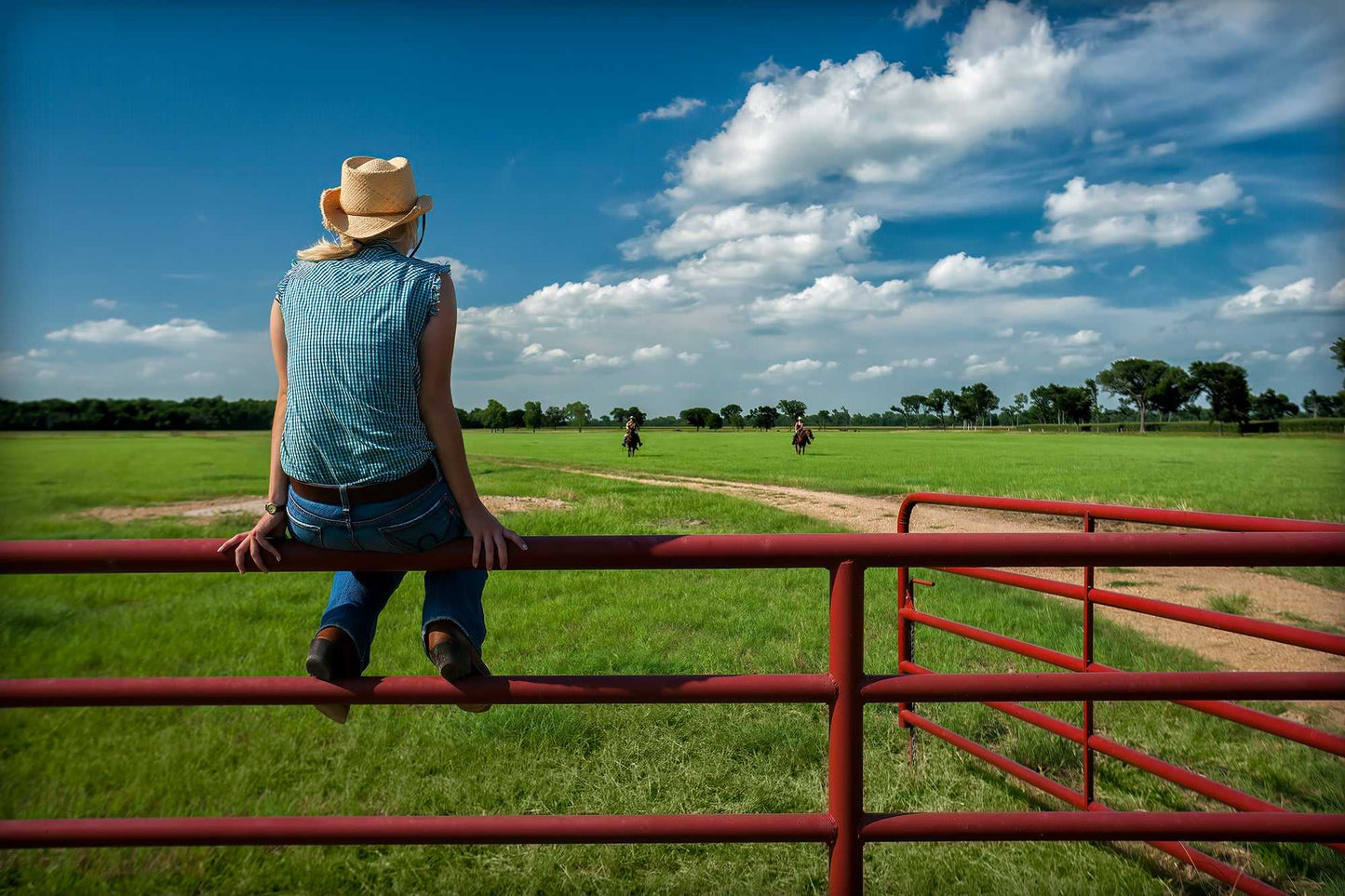 Coming Home: Waiting on the Fenceline at 44 Farms shows a cowgirl on a red fence watching cowboys ride in under a vivid Texas sky. A serene Western ranch scene.