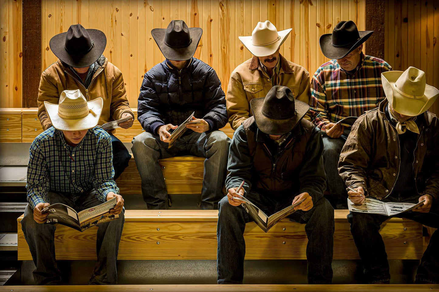 Before the Sale – Fine Art Ranch Photography print showing cowboys in hats seated on bleachers at 44 Farms, Texas, reading catalogs indoors.