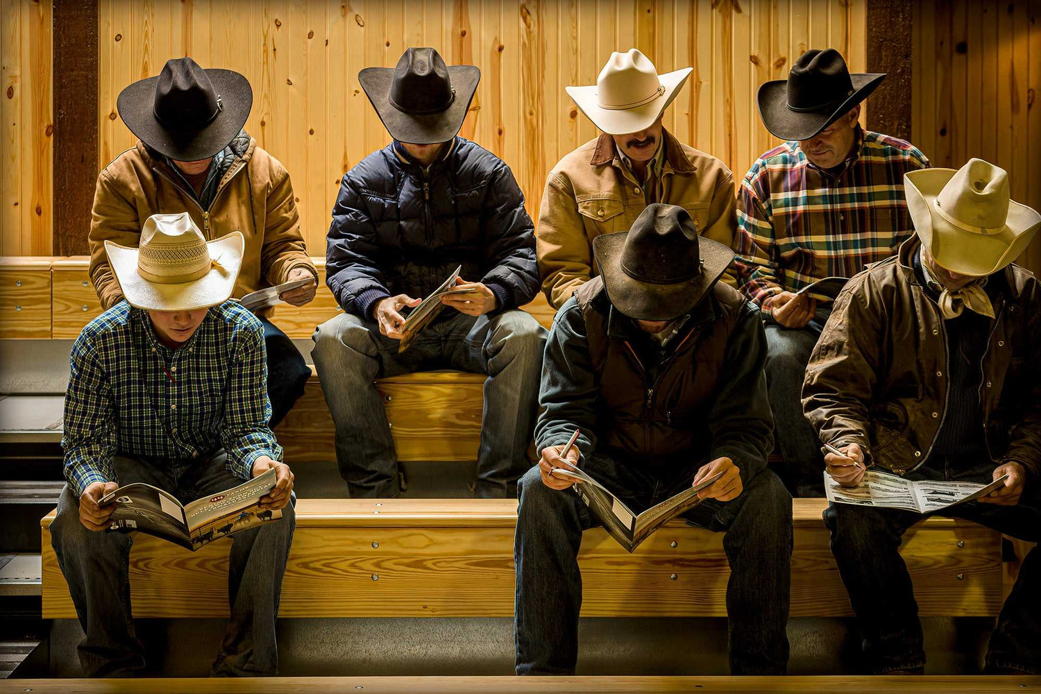 Before the Sale – Fine Art Ranch Photography print showing cowboys in hats seated on bleachers at 44 Farms, Texas, reading catalogs indoors.