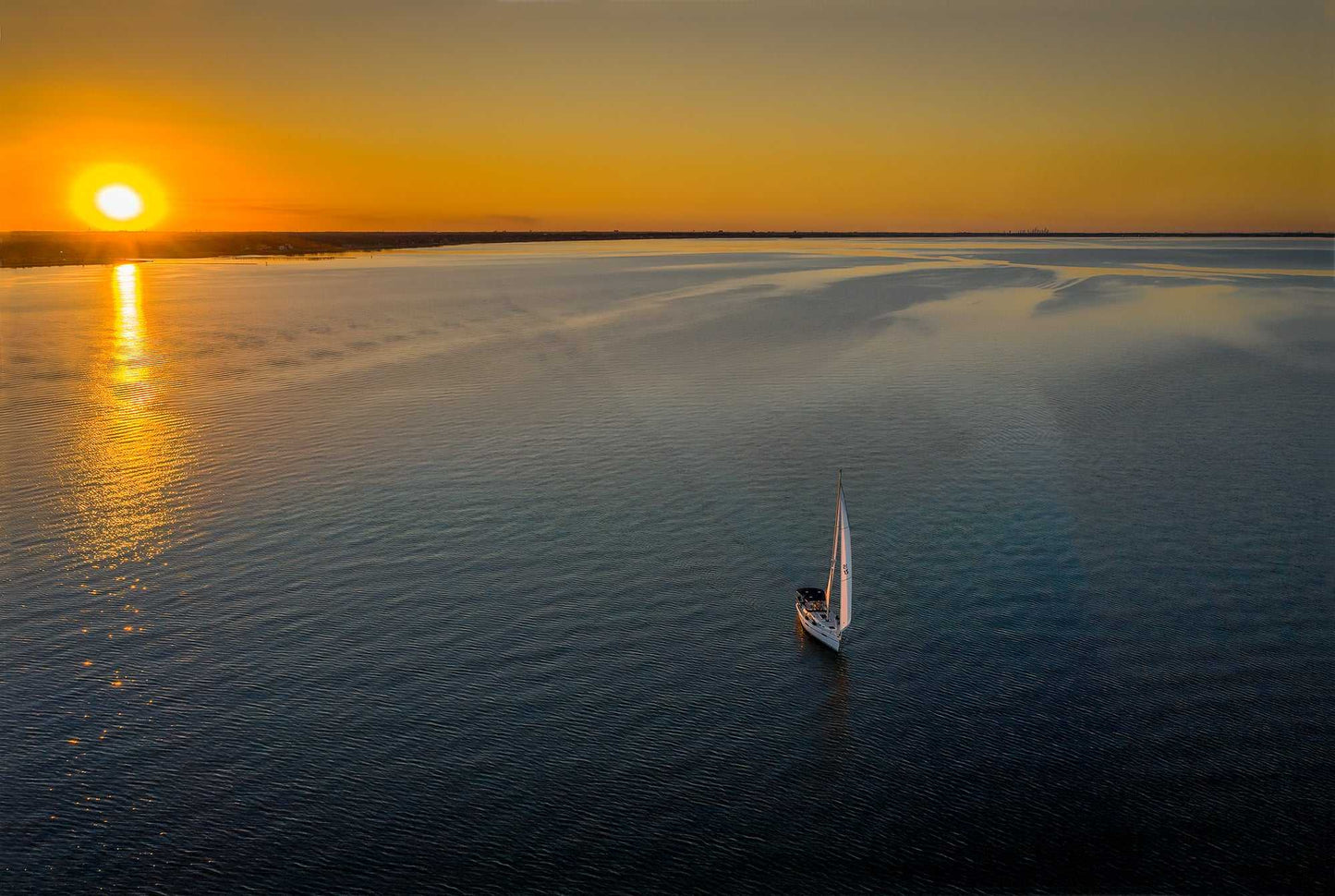 Sunset Sail – Fine Art Aerial Photograph Over Galveston Bay shows a sailboat gliding across calm waters at sunset. Fiery reflections light up the bay.