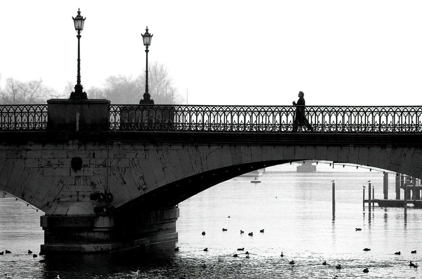 Zurich Series #1 – A Man on the Bridge, Zurich in Black and White – Fine Art Print by Michael Hart. A lone figure walks across a foggy bridge over the River Limmat in a monochrome cityscape.