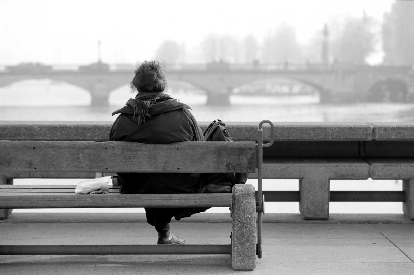 Zurich Series #6 – The Woman Overlooking the River Limmat by Michael Hart shows a solitary woman on a bench facing the Limmat in black and white. A serene fine art print capturing urban solitude in Zurich’s historic riverside.