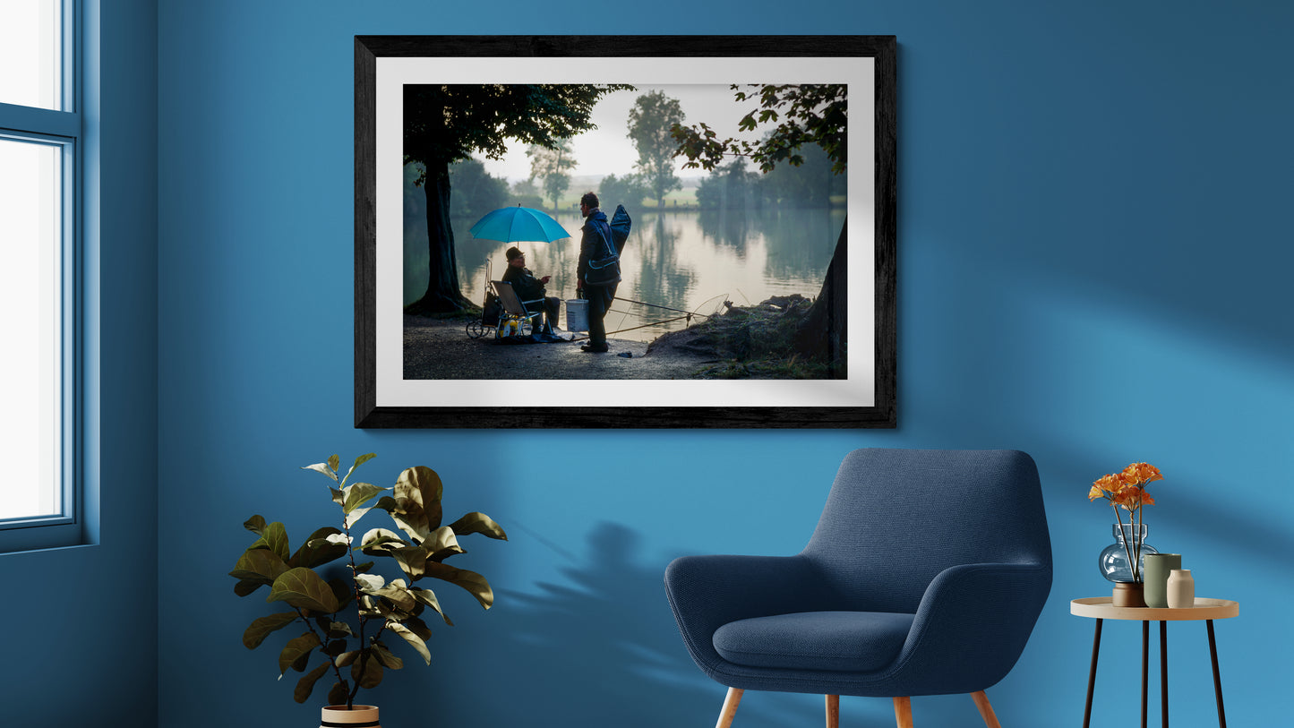 Framed fine art wall art featuring two fishermen with a blue umbrella displayed above a modern lounge chair against a blue wall with soft natural light and contemporary styling.