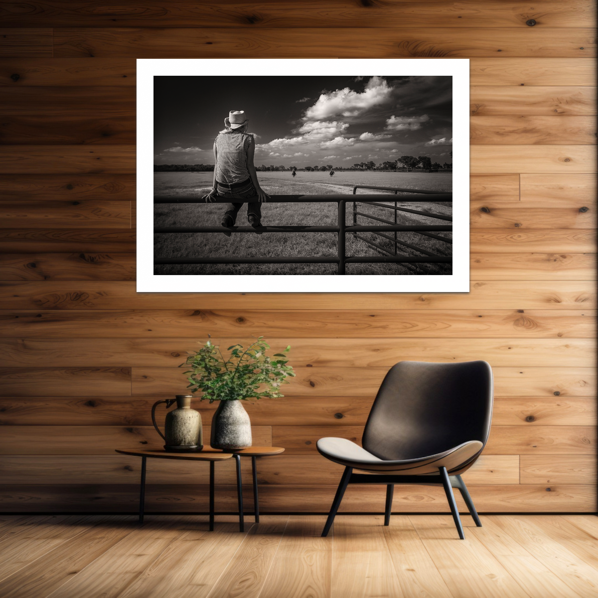 Framed black and white photo of ayoung woman sitting on a fence, displayed on a wooden wall above a chair and small table.