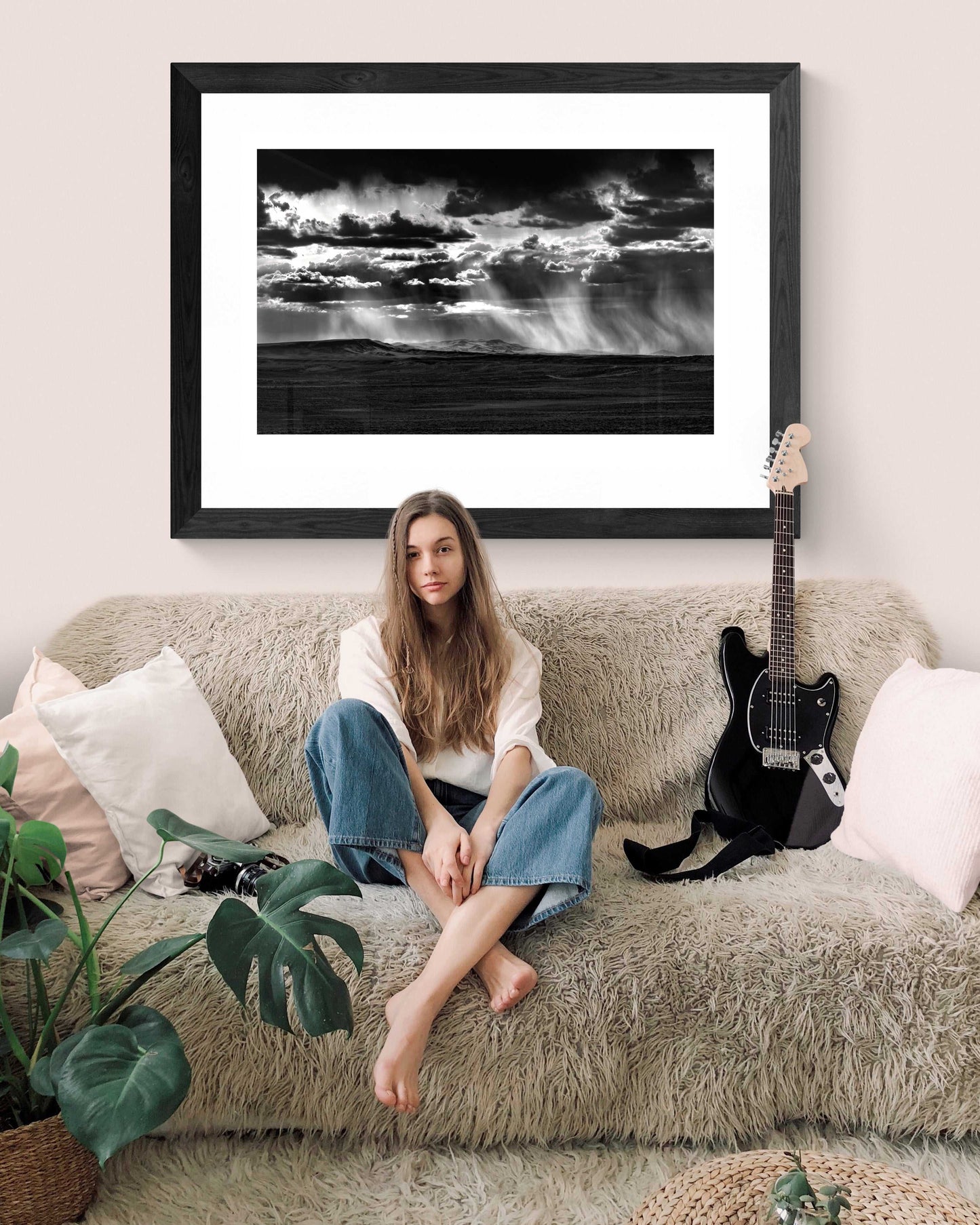 Wyoming Rainstorm – Backlit Virga Over the Plains framed black and white print displayed in a casual boho-style living room with a woman sitting on a plush sofa.