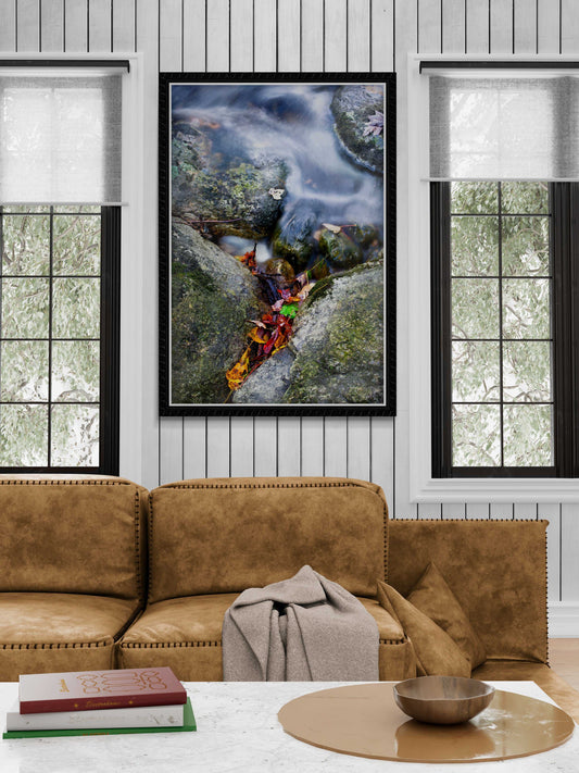 Framed First Frost fine art photograph displayed in a rustic living room, showcasing autumn leaves in a Maine stream above a tan leather sofa.