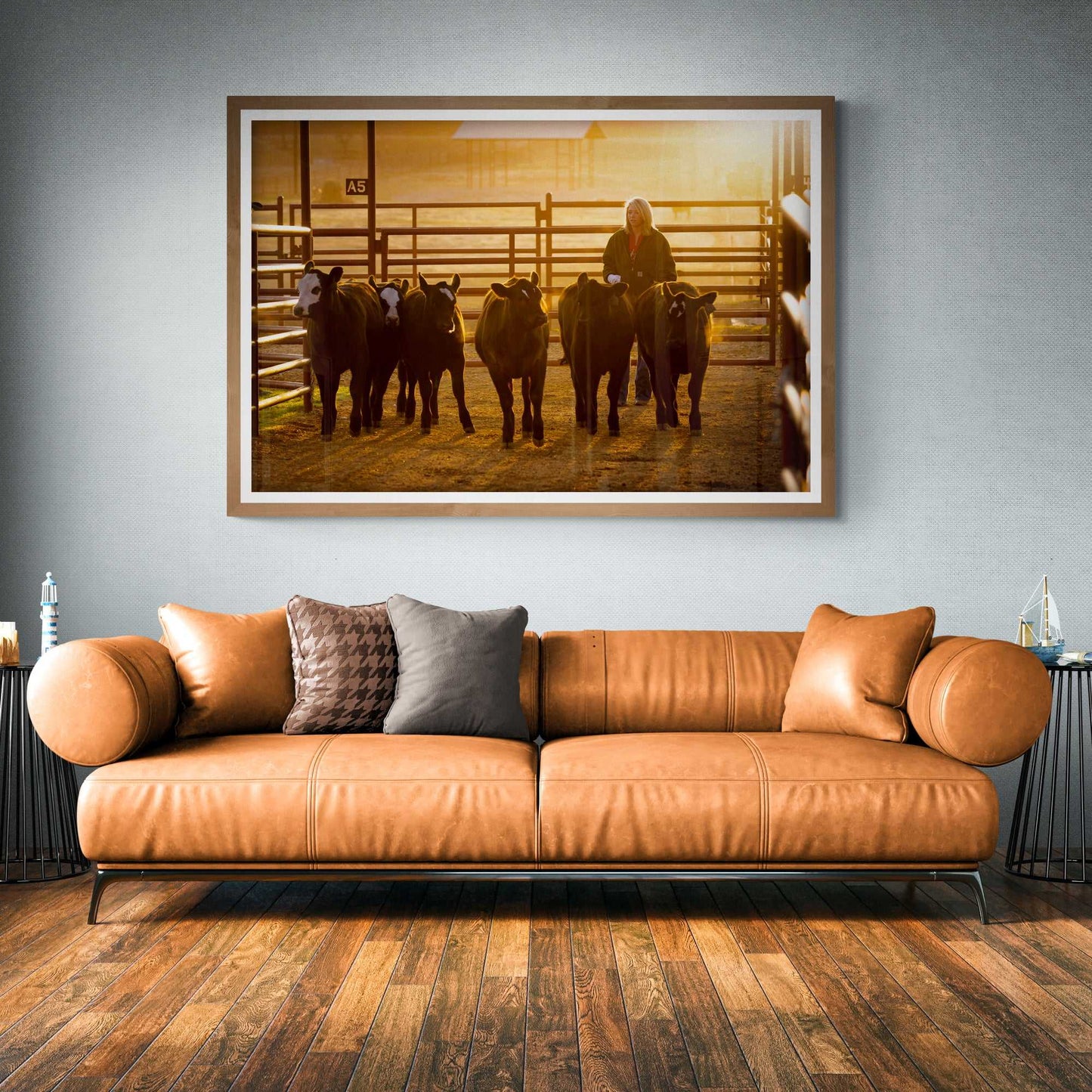 Calves at Daybreak: A Texas Ranch Morning in Golden Light fine art print displayed above a leather sofa. Features a female ranch hand guiding calves at sunrise.
