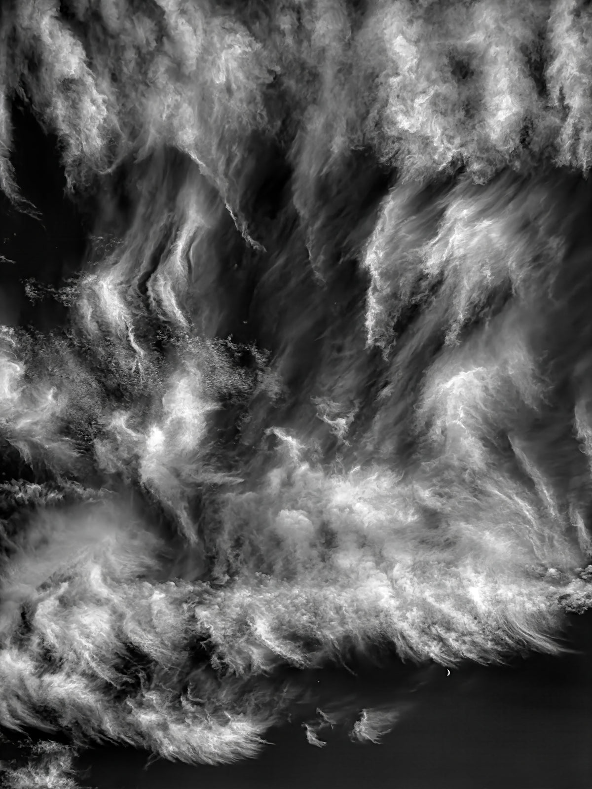 Dramatic black and white fine art photograph of swirling cirrus clouds with a small crescent moon beneath the clouds, titled The Moon Watches the Ride by Michael Hart.