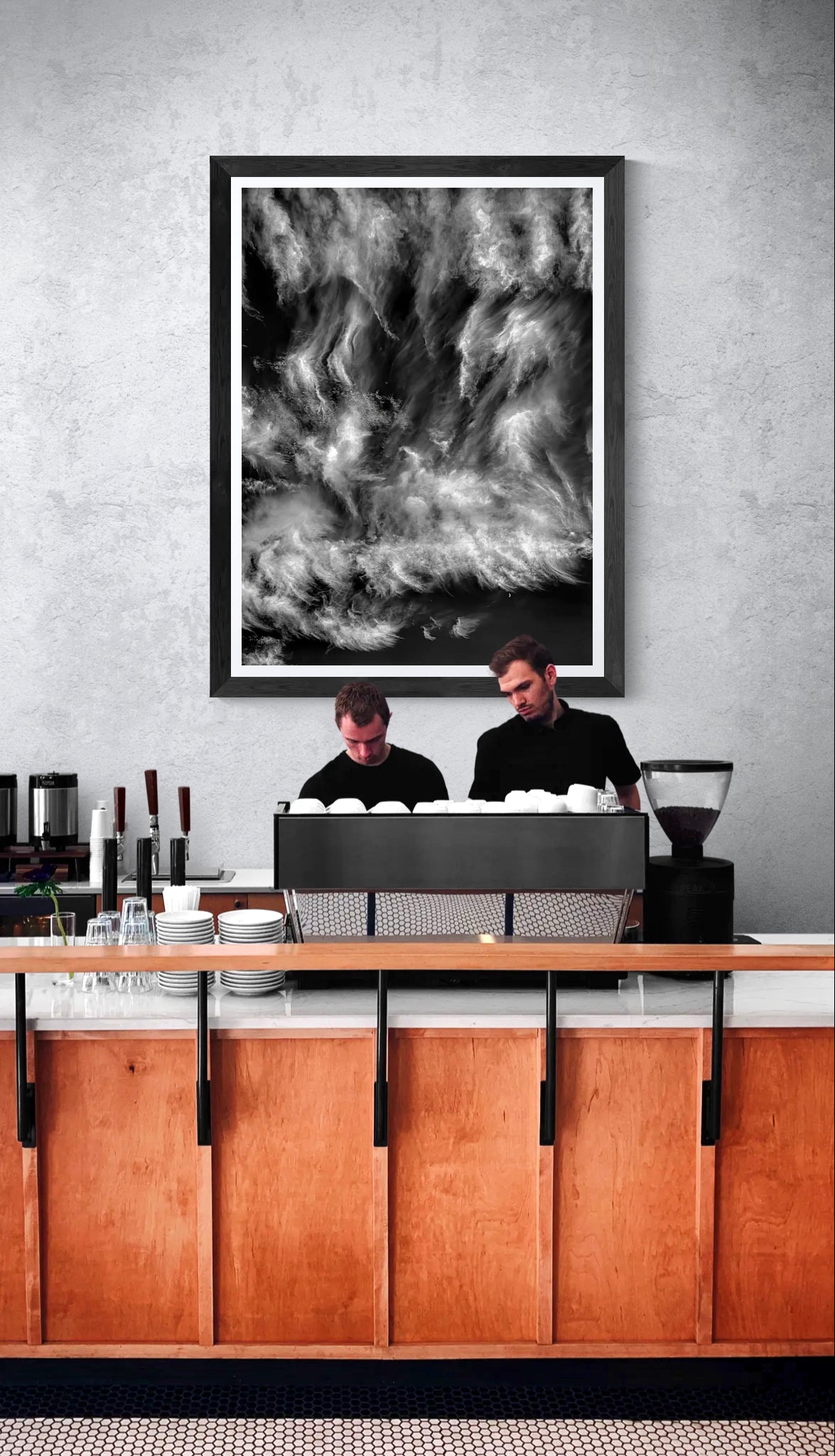 The Moon Watches the Ride, a limited edition black and white fine art photograph of swirling clouds and a crescent moon, displayed in a black frame above the counter of a contemporary coffee shop.