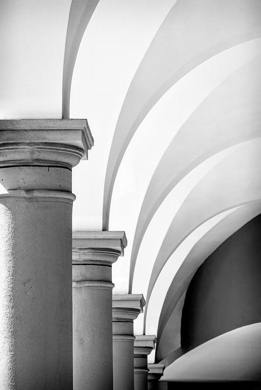 A black and white photo of some graceful arches in the Barberino outlet center in Italy. The graceful arches emanate from the top of the row of pillars and fan out vertically to our right.