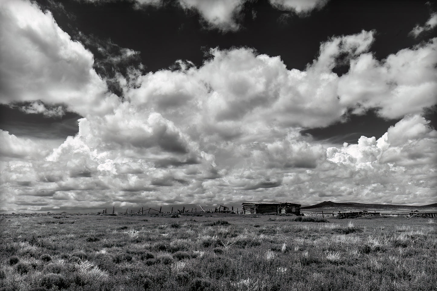 A brilliant and rich black and white image of the New Mexico landscape, it shows weathered ranch structures and fences under the billowing cumulus clouds of a summer afternoon.