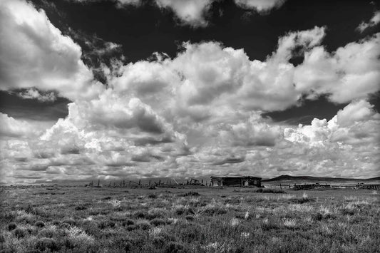 Abandoned Ranch — New Mexico black and white photograph of weathered structures beneath expansive cumulus clouds, a stark Western landscape defined by light and scale