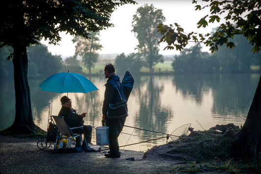 Two fishermen by Monrepos Lake in Germany, one seated beneath a blue umbrella, captured in soft evening light.