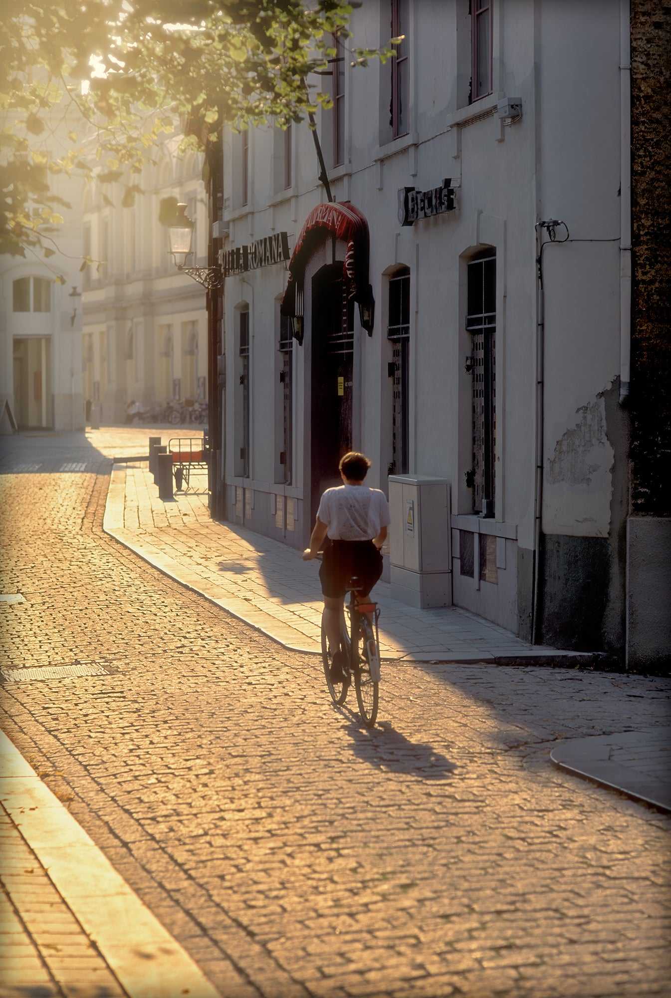 
Cyclist riding through golden light on a cobbled street in Bruges, Belgium, captured on Kodachrome film with long shadows.