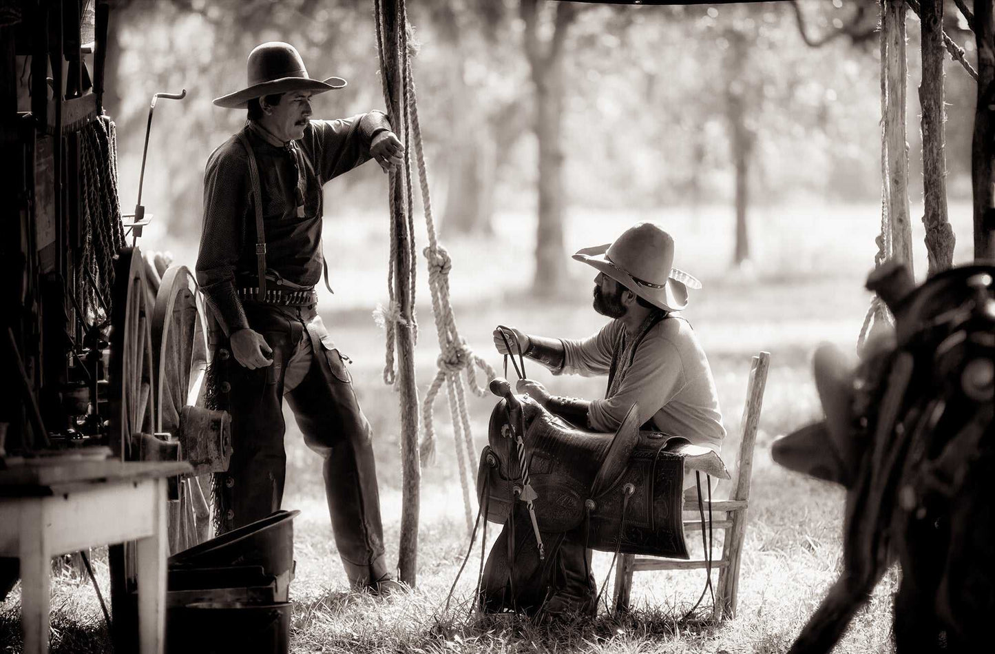 Two cowboys talking beside a chuckwagon during a Texas cook-off, surrounded by tack, rope, and camp gear in sepia tones.