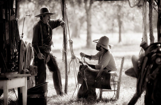 Two cowboys talking beside a chuckwagon during a Texas cook-off, surrounded by tack, rope, and camp gear in sepia tones.