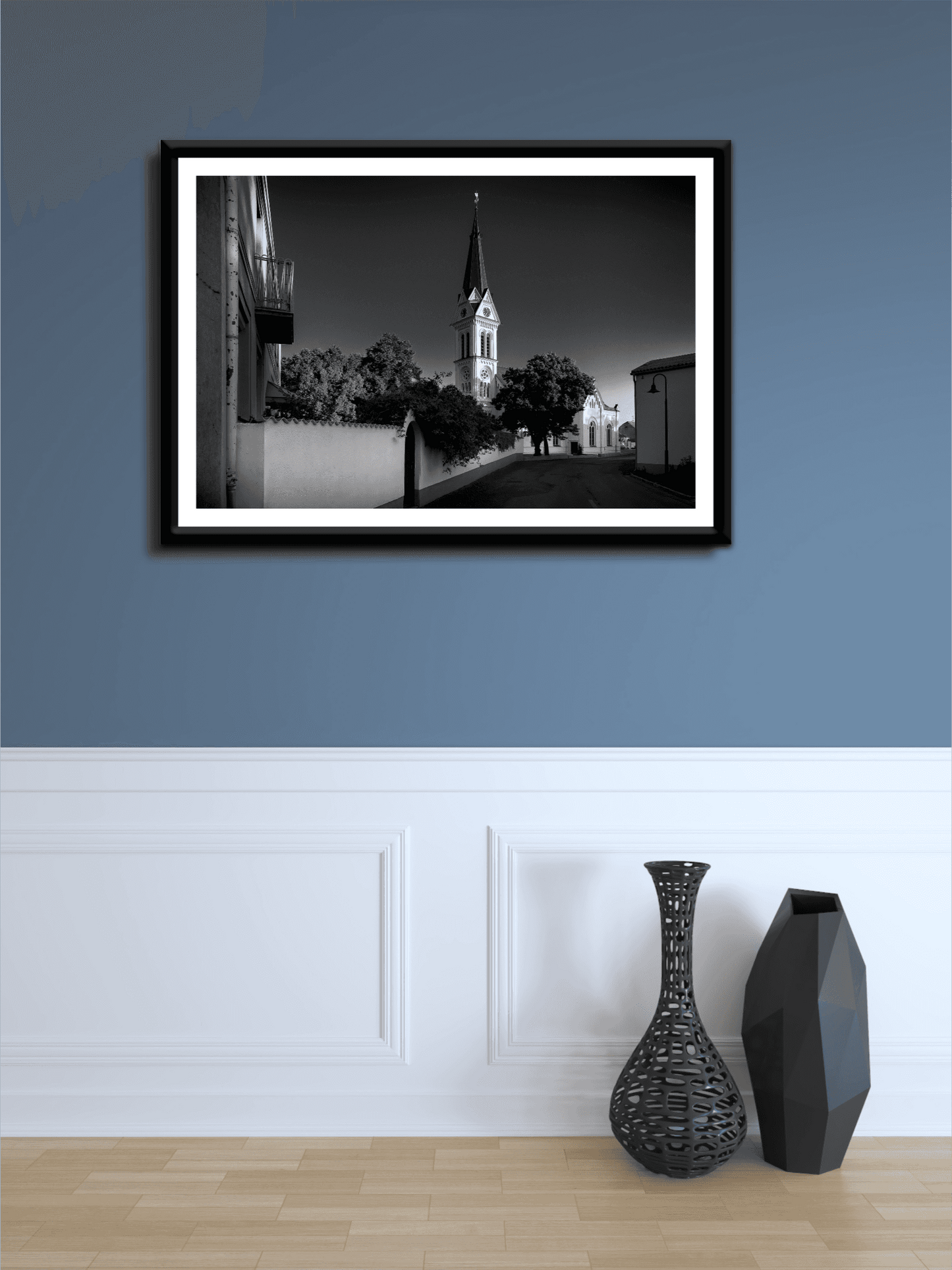A framed print of the black and white image of an Autrian Church and Steeple on a blue wall with white wainscoting.