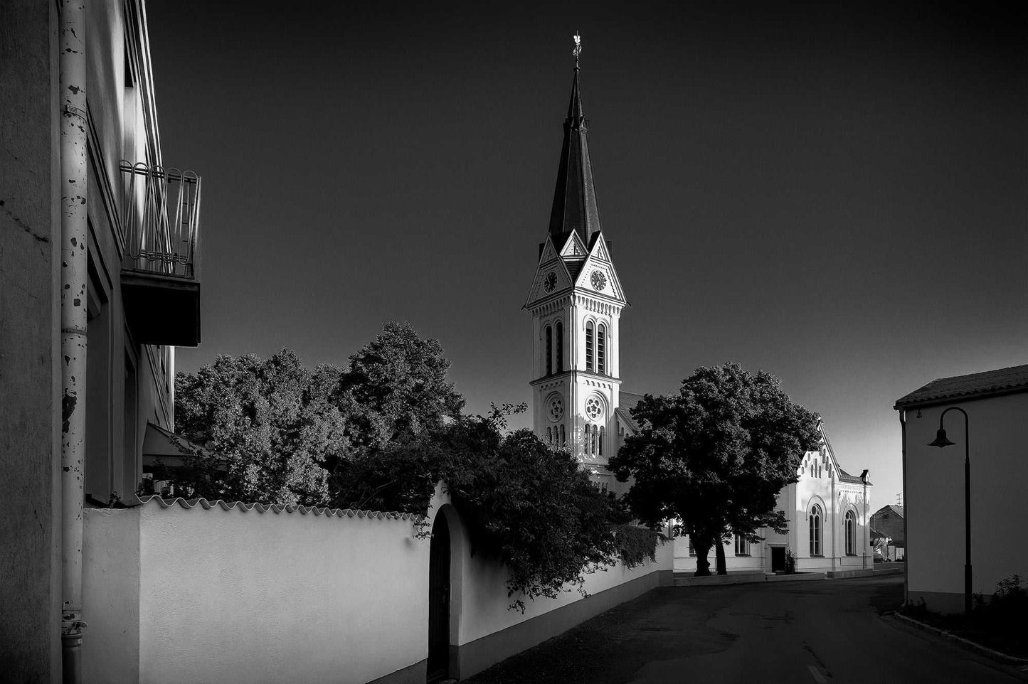 Church steeple in Neusiedl am See, Austria — black and white photograph of a village street leading to a pointed tower framed by trees and whitewashed buildings