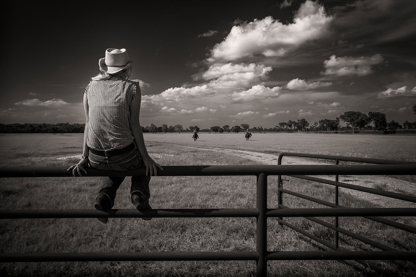Black and white photograph of a lone figure sitting on a fence watching riders return across a Texas ranch at 44 Farms.