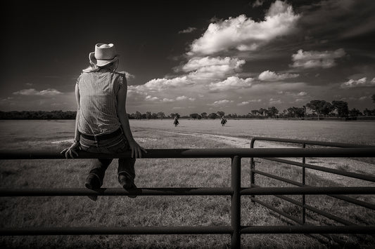 Black and white photograph of a lone figure sitting on a fence watching riders return across a Texas ranch at 44 Farms.