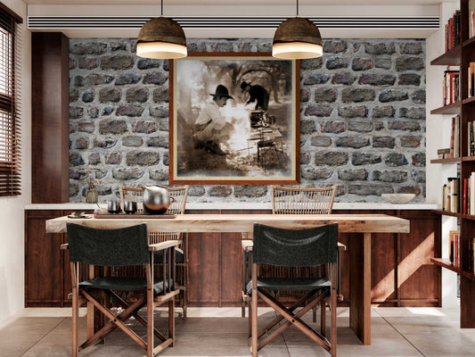 Framed Western heritage art print of chuckwagon cooking scene displayed in rustic dining room with stone wall, ideal for ranch-style home decor.