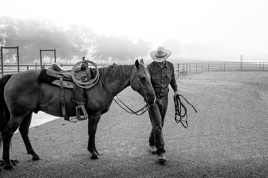 Black and white photograph of a cowboy leading a saddled horse through morning fog at 44 Farms in Texas