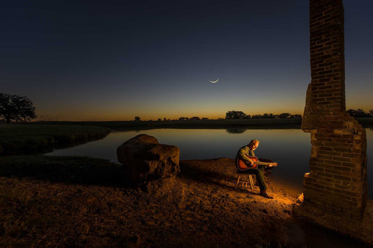 Musician playing guitar beside a fire at dusk on a Texas ranch at 44 Farms, with crescent moon over water