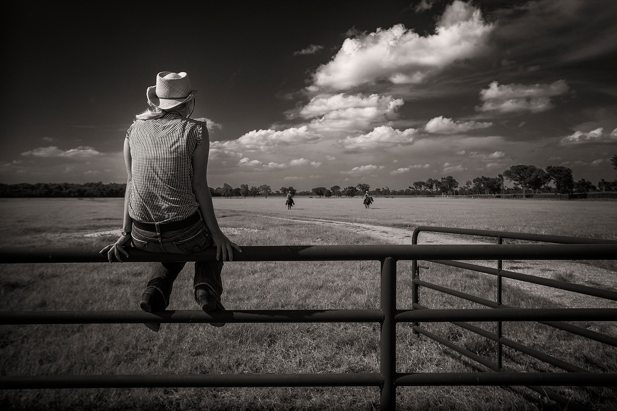 A young woman sits on a Texas ranch fence as the cowboys ride up at the end of the day.