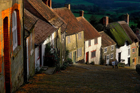 Fine art photograph of Gold Hill in Shaftesbury, Dorset, England, showing a steep cobbled street with historic cottages in warm afternoon light and a couple walking.