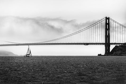 Black and white photograph of a sailboat beneath the Golden Gate Bridge, San Francisco, with fog and suspension cables
