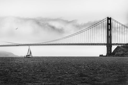 Black and white photograph of a sailboat beneath the Golden Gate Bridge, San Francisco, with fog and suspension cable