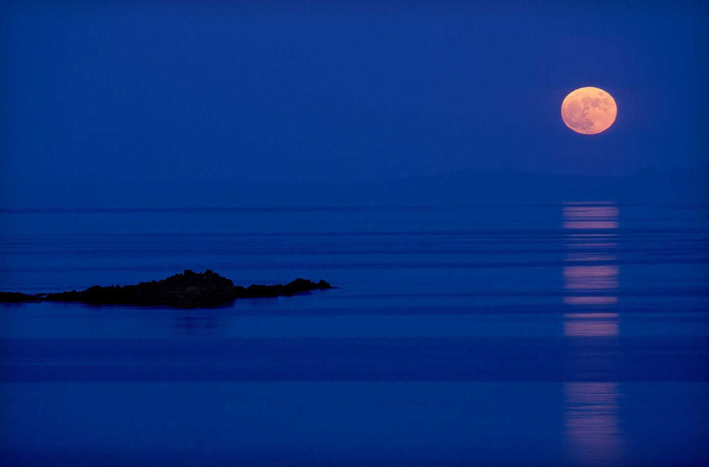 Full moon rising over calm Atlantic waters near the Isle of Islay, Scotland, with reflection stretching across the sea in blue twilight.