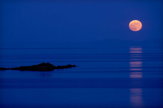 Full moon rising over calm Atlantic waters near the Isle of Islay, Scotland, with reflection stretching across the sea in blue twilight.