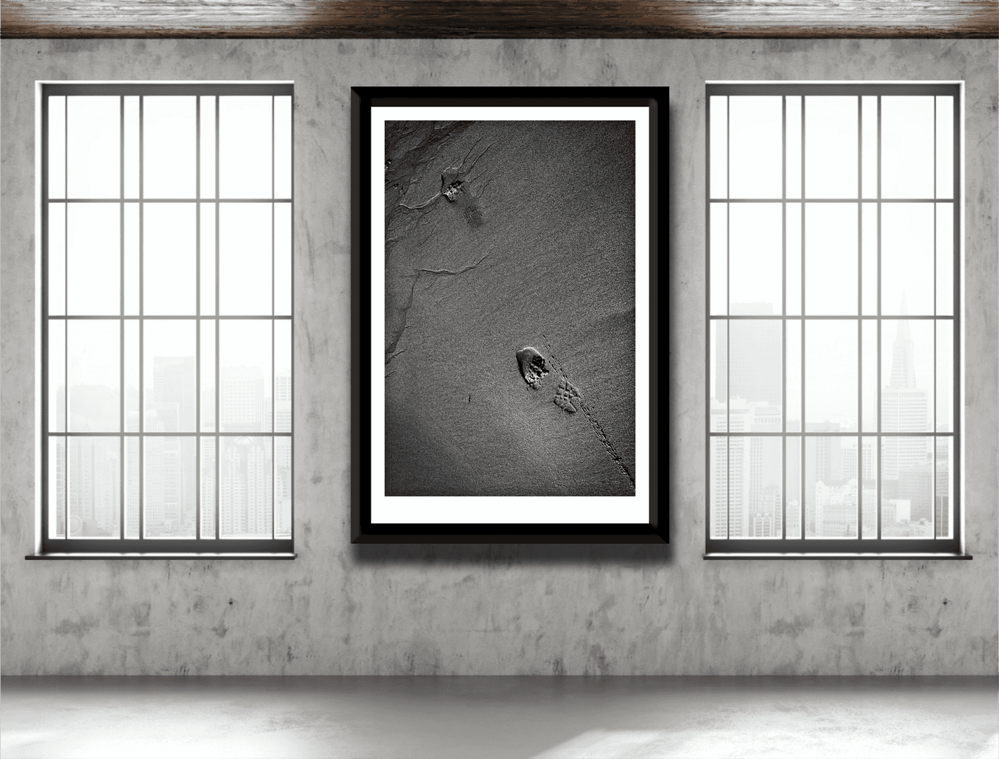 Large framed Traces #1 black and white sand photography with footprints, displayed in an industrial-style room with large windows.