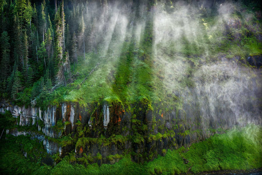 Sunlight streaming through mist at Mesa Falls in Idaho, illuminating moss-covered cliffs and forest in a lush, atmospheric landscape.