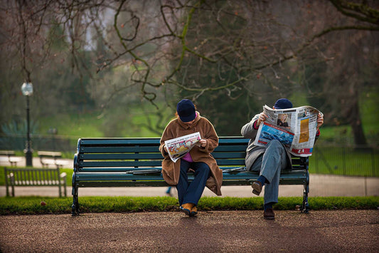 Two people reading newspapers on a bench in Hyde Park London, captured in soft morning light with a quiet, candid atmosphere.
