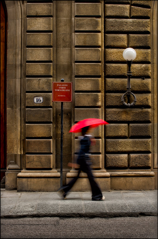 Woman with red umbrella walking past Palazzo Tornabuoni in Florence, Italy—street photography capturing motion against historic architecture.