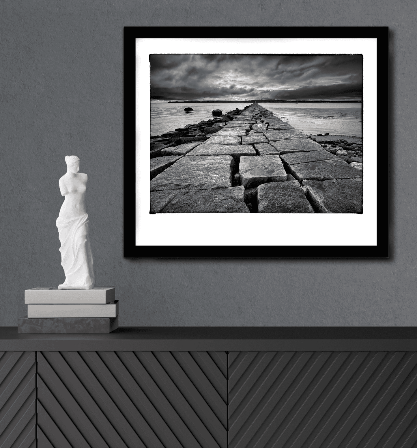 Breakwater, Rockland ME – Award-Winning Black & White iPhone Photo, Limited Edition Print framed on a modern gray wall above a console with a white sculpture. Dramatic granite jetty leads into a moody coastal horizon, ideal for fine art interior design.
