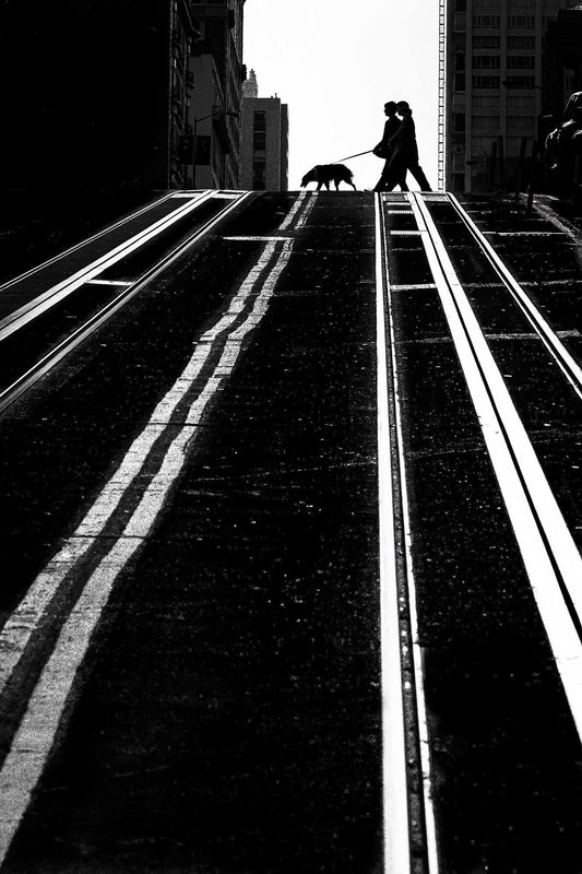 San Francisco Photography-Serendipitous Street Art print shows a silhouetted couple walking a dog across a steep hill with cable car tracks. Limited Edition black and white fine art by Michael Hart.