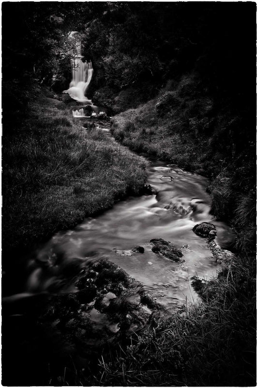 Black and white photograph of a flowing stream and small waterfall in Sutherland, Scotland, captured with long exposure to create smooth water against dark textured landscape