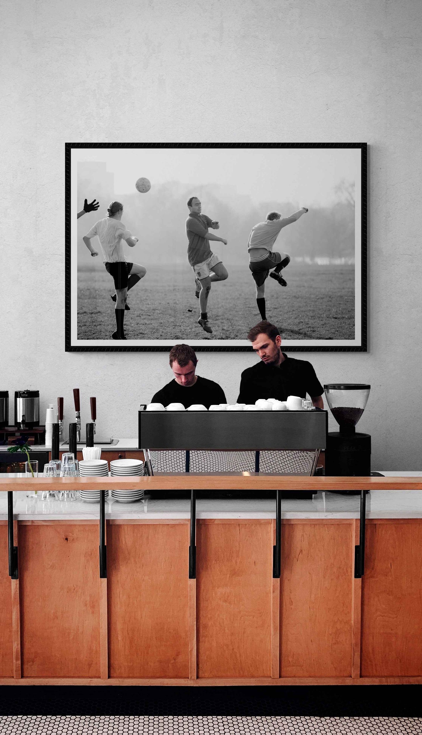 Black and white street photography print of three men playing football in Hyde Park, London on the wall behind baristas in a trendy coffee shop.