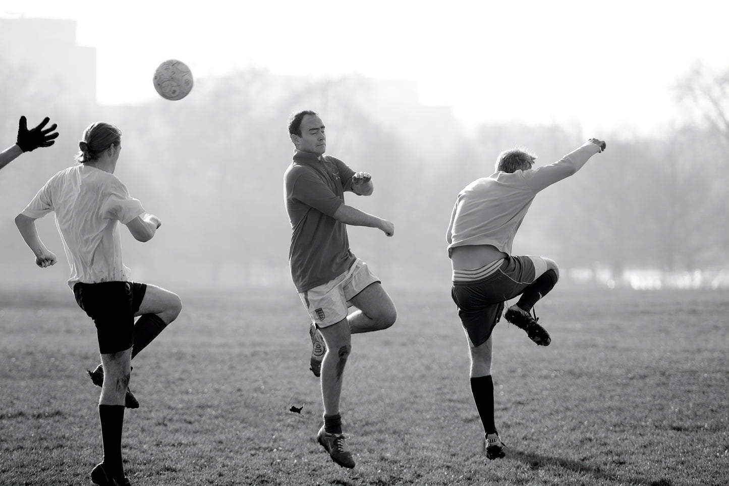 Black and white photograph of men playing football in Hyde Park, London, captured mid-kick with ball suspended and limbs in motion.