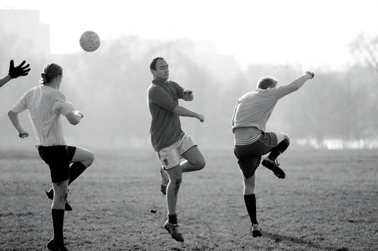 Black and white photograph of men playing football in Hyde Park, London, captured mid-kick with ball suspended and limbs in motion.