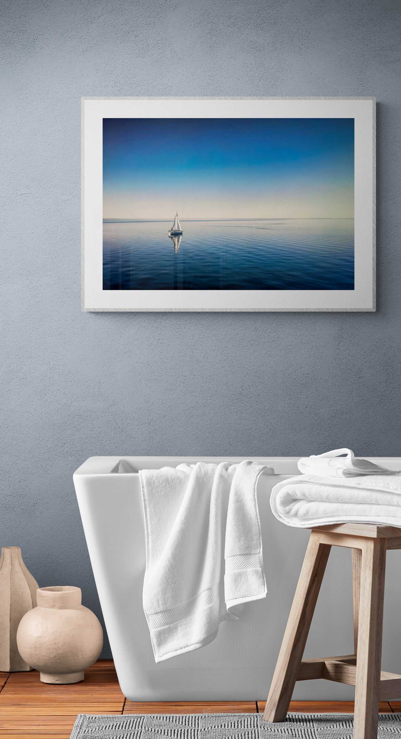 The Calm – Sailboat on Calm Blue Galveston Bay print adds peaceful seascape charm to a minimalist bathroom with soft blue-gray walls and white decor.