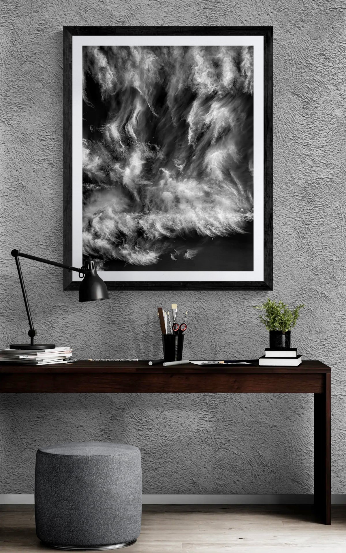 Black and white fine art photograph of dramatic swirling clouds and a small crescent moon displayed in a black frame above a dark wood desk against a textured gray wall in a contemporary home office.