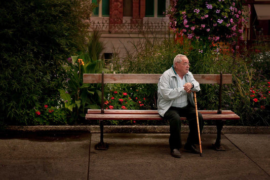 Elderly man seated on a bench with a cane beneath flowers in Troyes, France, captured as a quiet street photography moment.