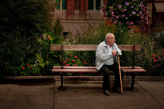 Elderly man seated on a bench with a cane beneath flowers in Troyes, France, captured as a quiet street photography moment.