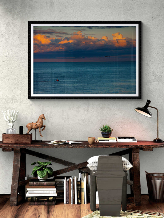 Framed print of The Race – Small Boat and Birds in a New England Coastal Sunset displayed above a rustic desk, ideal for coastal-themed interior design.