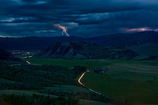 Lightning storm over Jackson Hole Wyoming with car light trails winding through valley at dusk